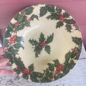 May include: A white ceramic bowl with a green and red holly leaf design. The bowl is round and has a slightly raised rim.