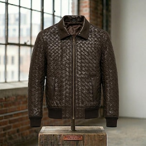 May include: A dark brown jacket featuring a woven leather pattern across the body and sleeves. The jacket includes a solid brown collar, a central metal zipper, two side pockets, and dark brown ribbed cuffs and hem at the bottom edge.