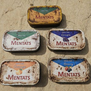 May include: Five rectangular metal tins with the word "MENTATS" in a distressed font. Each tin features a different flavour label: "Grape", "Orange", "Berry", and "Mentats". The tins have a weathered, aged appearance, suggesting they are vintage.