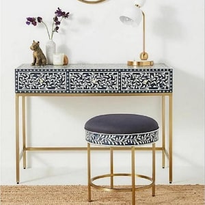 Puede incluir: Un juego de tocador con estructura de metal dorado, con un diseño floral en azul oscuro y blanco. El conjunto incluye un taburete a juego y un espejo redondo. Una lámpara blanca y objetos decorativos están sobre el tocador.