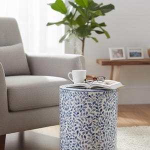 May include: A cylindrical side table with a white and blue leaf pattern. A white mug, glasses, and a book rest on top. The table is next to a gray armchair and a green plant.