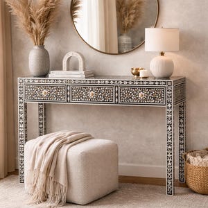 May include: A decorative console table with three drawers, featuring a black and white floral inlay design. A round mirror hangs above the table, reflecting the room. A beige ottoman with a fringed throw blanket sits in front of the table. A lamp and vase are on the table.