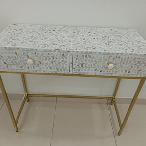 Puede incluir: Una consola blanca con dos cajones, adornada con un diseño floral. La mesa tiene una estructura de metal dorado y tiradores blancos en forma de flor. Sus dimensiones son de aproximadamente 102 cm de ancho, 38 cm de profundidad y 76 cm de alto.