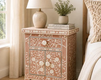May include: A rectangular bedside table with a dusty rose finish and white floral inlay patterns. It features one top drawer and two lower cabinet doors, each equipped with white flower-shaped knobs. The table has a flat top surface.