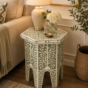 May include: A hexagonal side table featuring a sage green surface with intricate white floral and vine inlay patterns. The table has six straight legs with arched cutouts at the base, creating a structured silhouette with a flat top surface.