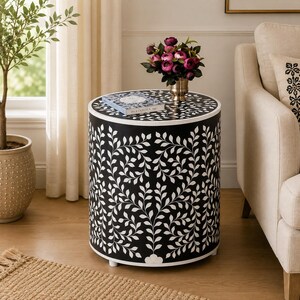 May include: A cylindrical side table featuring a black base with a white inlaid leaf pattern. The top surface holds a book titled The Kinfolk and a gold-toned vase containing a bouquet of dark pink flowers. The table stands on small feet.