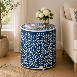May include: A cylindrical side table featuring a deep blue finish with a repeating white vine and leaf pattern. The top surface holds a book titled The Kinfolk Home and a silver vase containing a bouquet of white hydrangea flowers.