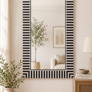 Pode incluir: Um espelho de parede retangular com uma moldura de padrão listrado preto e branco repetitivo. A moldura consiste em barras pretas verticais e horizontais que se alternam com espaços brancos, criando um design de borda geométrico.