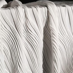 Könnte beinhalten: Nahaufnahme eines weißen Stoffes mit einem plissierten Design. Der Stoff ist drapiert und erzeugt weiche Falten und Schatten. Die Textur scheint gerippt zu sein, mit vertikalen Linien, die über das Material verlaufen. Der Gesamteffekt ist sauber und elegant.