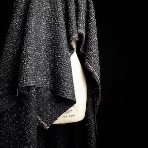 May include: A dark charcoal gray woven fabric with a textured, speckled appearance draped over a beige dress form. The fabric features a loose, open weave with visible light gray flecks throughout and raw, unfinished edges along the hemline.