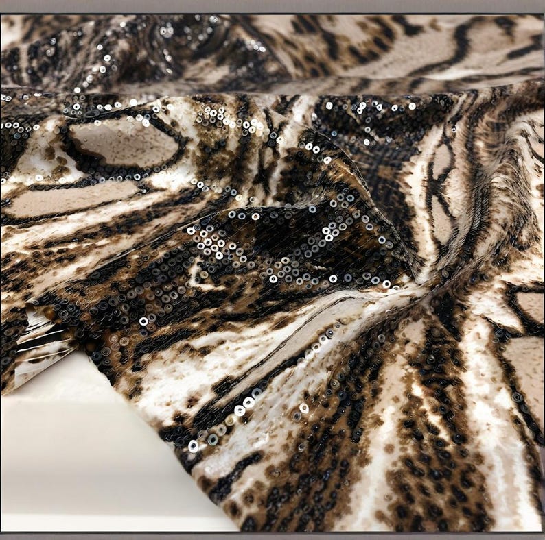 May include: A close-up shot of a fabric featuring a swirling pattern in brown, tan, and black, adorned with sparkling sequins. The material has a textured look, with folds and creases adding depth. The overall effect is luxurious and visually appealing.