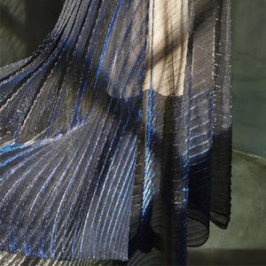Puede incluir: Una tela fluida y transparente con una combinación de colores azul oscuro y negro. La tela presenta rayas verticales con un brillo metálico, creando un efecto brillante. El material parece ligero y adecuado para drapeados.