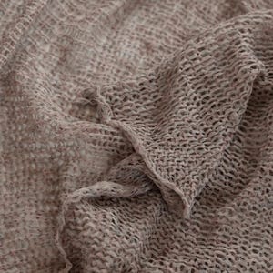 May include: Close-up of a loosely woven, light brown fabric. The material has a textured, open weave pattern, creating a delicate, airy appearance. The fabric is draped, with soft folds and subtle shadows, suggesting a lightweight textile.