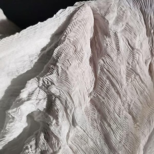 May include: Close-up of a white, textured fabric with a crinkled, wavy pattern. The fabric appears soft and lightweight, with subtle shadows adding depth to the folds. The overall effect is delicate and airy, suggesting a scarf or shawl.