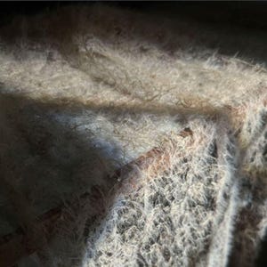 May include: Close-up of a textured, light-colored fabric with a dense, fuzzy appearance. The fabric has a mix of light and dark areas, with subtle variations in color and texture. The image captures the intricate details of the material.