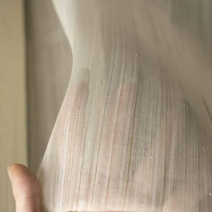 May include: Close-up of a sheer, off-white fabric with a vertical striped pattern. The fabric is held up, revealing the texture and translucence. The material appears delicate and lightweight, possibly for curtains or clothing.