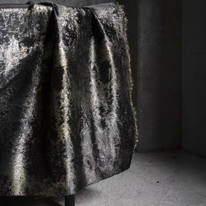 Puede incluir: Tela negra con un patr&oacute;n dorado met&aacute;lico drapeada sobre una superficie oscura. La tela tiene una apariencia texturizada con flecos en el borde. La est&eacute;tica general es lujosa y elegante.