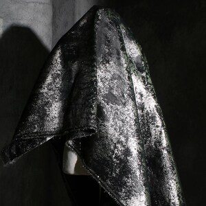 Puede incluir: Tela negra drapeada con un aspecto met&aacute;lico moteado. La tela presenta reflejos plateados y verdes, creando un efecto texturizado y brillante. La tela est&aacute; drapeada sobre una forma invisible, sugiriendo una prenda o textil decorativo.
