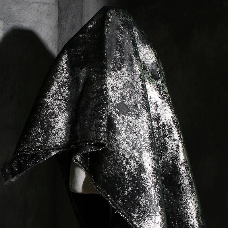 Puede incluir: Tela negra drapeada con un aspecto met&aacute;lico moteado. La tela presenta reflejos plateados y verdes, creando un efecto texturizado y brillante. La tela est&aacute; drapeada sobre una forma invisible, sugiriendo una prenda o textil decorativo.