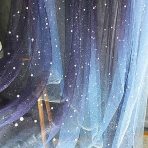 May include: A flowing sheer fabric with a colour gradient from dark indigo to light blue. The fabric is decorated with scattered white pearl-like embellishments, creating a starry night effect. The fabric is likely used for decorative purposes.