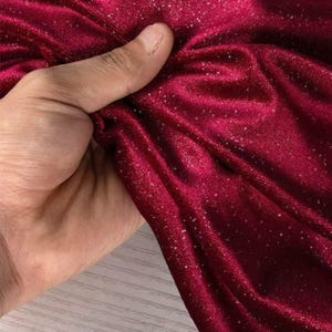 May include: Close-up of a deep red fabric with a velvety texture and sparkling glitter accents. The fabric is being held, showcasing its drape and sheen. The material appears soft and luxurious, suitable for apparel or decorative projects.