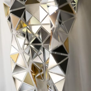 May include: Close-up of a garment with a geometric design. The surface is covered in small, reflective triangles in silver, gold, black, and white, creating a mosaic effect. The triangles are arranged in a repeating pattern.
