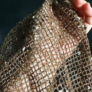 Pode incluir: Close-up de um tecido dourado de trama aberta com um padrão de grade. O tecido tem um brilho metálico e pequenas manchas brancas. O material é segurado, revelando a pele por baixo. A textura geral parece delicada e arejada.