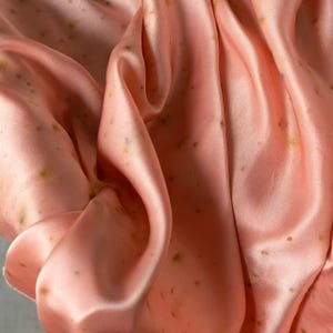 Könnte beinhalten: Nahaufnahme eines fließenden, pfirsichfarbenen Stoffes mit dezentem Glanz. Das Material ist in weichen Falten drapiert, wodurch Schatten und Lichtreflexe entstehen. Kleine, unregelmäßige, dunkelgrüne Punkte sind über die Oberfläche verstreut.