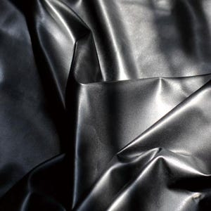 Op de afbeelding: Close-up van een zwarte, glanzende stof met een gladde, leerachtige uitstraling. Het materiaal is gevouwen en gedrapeerd, waardoor schaduwen en highlights ontstaan die de textuur en glans benadrukken. De stof lijkt effen van kleur.