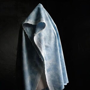 Op de afbeelding: Een lichtblauwe cape met capuchon met een getextureerd oppervlak. De cape is gedrapeerd en onthult een glimp van een lichtere stof eronder. De capuchon is puntig en de randen zijn afgewerkt met een witte bies. De achtergrond is zwart.