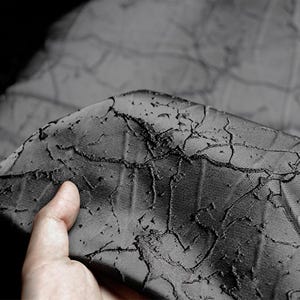 以下が含まれることがあります： ひび割れた模様の、質感のある濃いグレーの生地のクローズアップ。生地は隆起した3次元効果があり、独特の視覚的テクスチャを作り出しています。素材は柔らかくしなやかで、わずかな光沢があります。