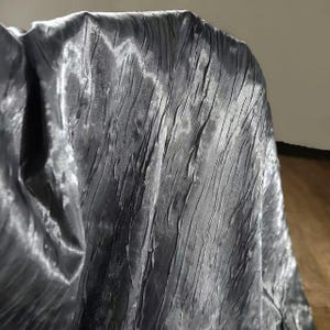 May include: Close-up of a draped, textured fabric in a dark gray color. The fabric has a crinkled, wrinkled appearance with vertical lines and a subtle sheen. The material appears to be soft and flowing, with highlights and shadows.