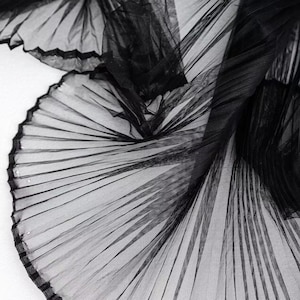 Può includere: Primo piano di tessuto nero plissettato, che mostra pieghe e trame intricate. Il materiale trasparente crea un effetto a strati, con luce e ombra che giocano sulla superficie. Il tessuto è probabilmente utilizzato per scopi di moda o decorativi.