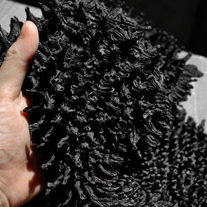 May include: Close-up of a pile of black, textured fabric. The fabric has a dense, layered appearance with a rough, uneven surface. The image is in black and white, highlighting the intricate details and shadows of the material.
