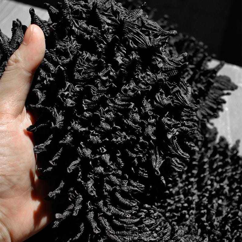 May include: Close-up of a pile of black, textured fabric. The fabric has a dense, layered appearance with a rough, uneven surface. The image is in black and white, highlighting the intricate details and shadows of the material.