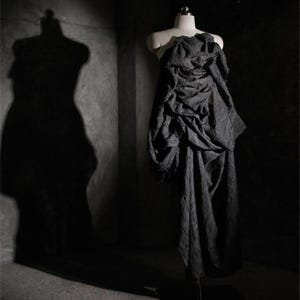May include: A dark gray, draped dress on a mannequin. The dress has a textured appearance and a flowing design. The image is set in a dimly lit space, with the shadow of a dress form visible on the wall.
