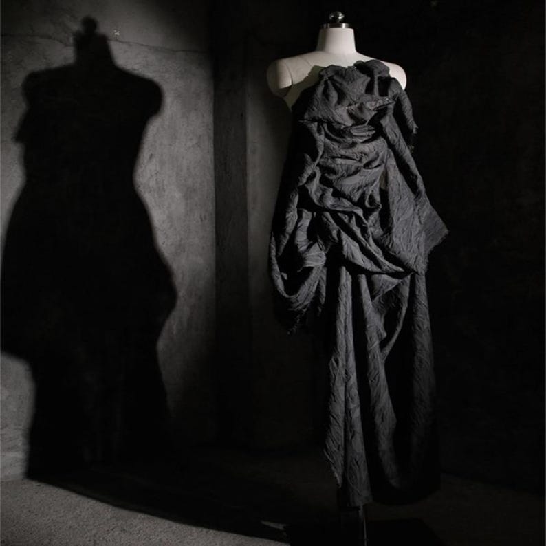 May include: A dark gray, draped dress on a mannequin. The dress has a textured appearance and a flowing design. The image is set in a dimly lit space, with the shadow of a dress form visible on the wall.