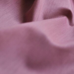 May include: Close-up of a folded fabric in a dusty rose color. The fabric has a subtle sheen and a textured surface, with visible folds and shadows creating depth. The image highlights the material's smooth appearance.