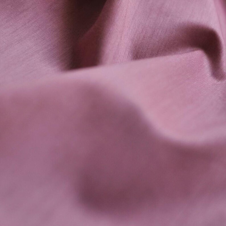 May include: Close-up of a folded fabric in a dusty rose color. The fabric has a subtle sheen and a textured surface, with visible folds and shadows creating depth. The image highlights the material's smooth appearance.