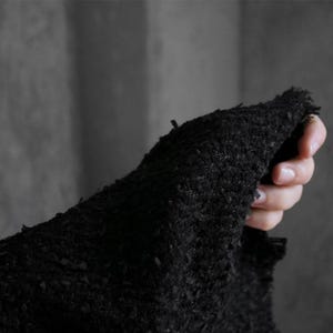 May include: Close-up of a textured black fabric. The fabric appears to be thick and rough, with a frayed edge. The image is in soft focus, with the fabric taking up most of the frame. The background is a muted gray.