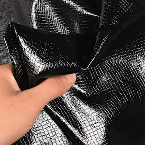 Może przedstawiać: Zbliżenie czarnego, teksturowanego materiału z błyszczącym wykończeniem. Materiał ma wzór przypominający łuski i jest trzymany, pokazując jego elastyczność i połysk. Tkanina wydaje się być skórą lub materiałem skóropodobnym.