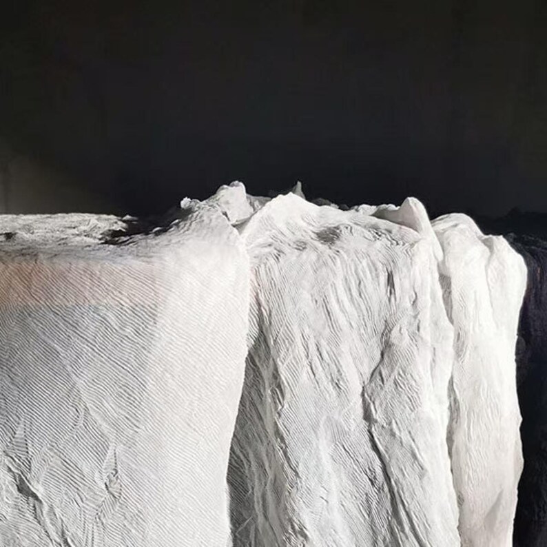 May include: Close-up of several rolls of white fabric with a textured, crinkled appearance. The fabric is arranged in a layered formation, with a dark background. The image highlights the texture and folds of the material.