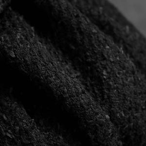 May include: Close-up of a textured, black fabric with visible folds and a slightly rough surface. The material appears to be thick and opaque, with subtle variations in tone, creating depth and shadow.