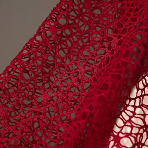 May include: Close-up of a vibrant red, open-weave fabric. The material has a delicate, net-like pattern with numerous small holes, creating a textured surface. The fabric is draped, showing folds and shadows, and appears soft and flexible.