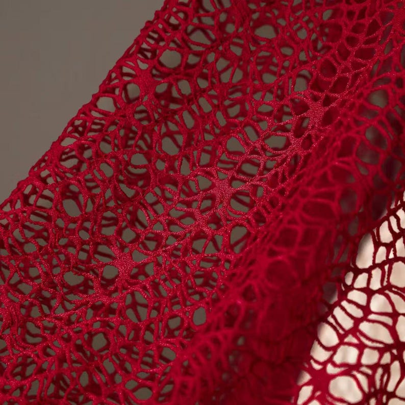 May include: Close-up of a vibrant red, open-weave fabric. The material has a delicate, net-like pattern with numerous small holes, creating a textured surface. The fabric is draped, showing folds and shadows, and appears soft and flexible.