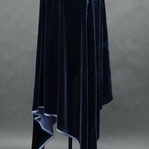 May include: A navy blue velvet cape with a draped design. The cape has a soft, luxurious texture and a contrasting white trim along the edges. The garment is displayed on a black stand against a gray background.