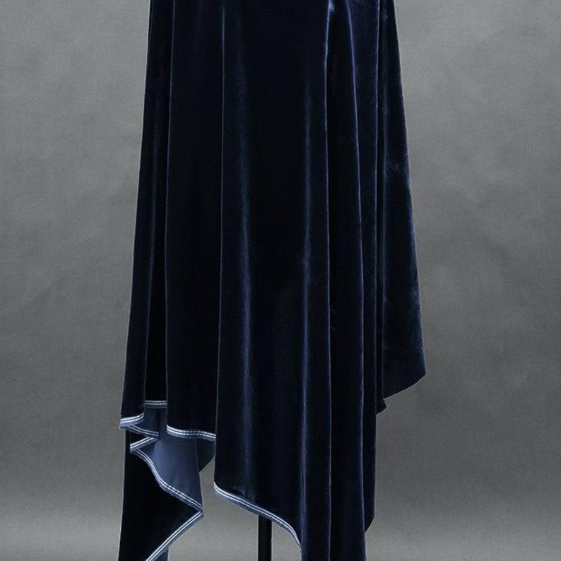 May include: A navy blue velvet cape with a draped design. The cape has a soft, luxurious texture and a contrasting white trim along the edges. The garment is displayed on a black stand against a gray background.