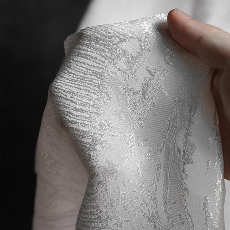 K&ouml;nnte beinhalten: Nahaufnahme eines wei&szlig;en Stoffes mit einem strukturierten, wirbelnden Muster. Der Stoff hat einen subtilen Glanz und ein detailliertes Design mit erhabenen Texturbereichen. Das Material wird gehalten und zeigt seinen Fall und seine Qualit&auml;t.