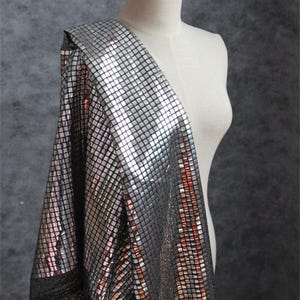 May include: A silver fabric featuring a dense grid of small, reflective square tiles. The material has a metallic, mirror-like finish that catches light across its surface, creating a shimmering effect. The fabric is draped over a mannequin form.