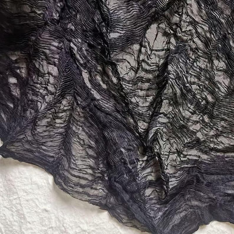 May include: Close-up of a dark gray, textured fabric with a crinkled, layered appearance. The fabric has a flowing, wave-like pattern, with lighter gray areas interspersed throughout. The material appears to be lightweight and semi-sheer, with a soft, draping quality.